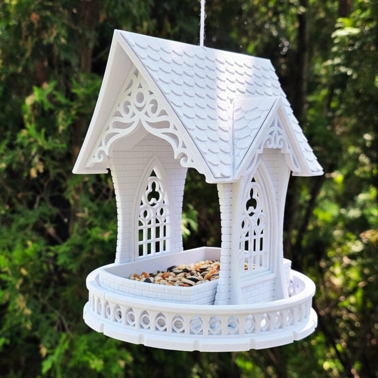 A white decorative hanging bird feeder shaped like a small pavilion or gazebo, suspended by a string outdoors against a blurred green foliage background.