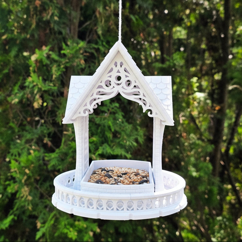 A white decorative hanging bird feeder with an ornate gabled roof and circular tray, suspended outdoors against a blurred green foliage background.