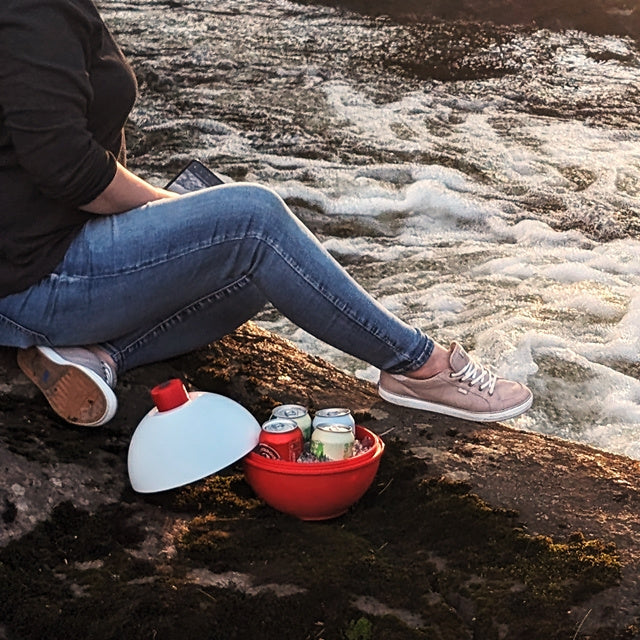 Person sitting by a river with a red container and bottles on a rock