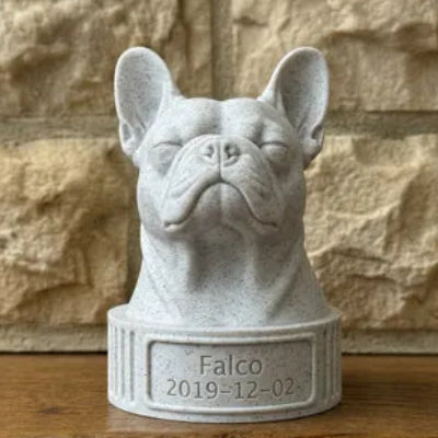 White stone sculpture of a french bulldog head with 'Falco' and a date engraved, on a wooden surface with a stone wall background.
