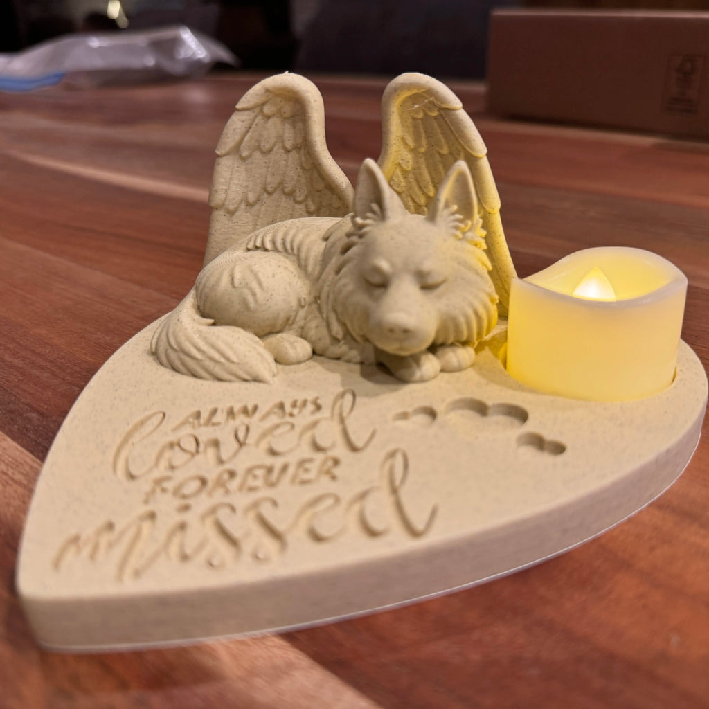 Memorial sculpture of a German shepherd with angel wings and a candle, engraved with 'Always Loved Forever Missed' on a wooden surface.