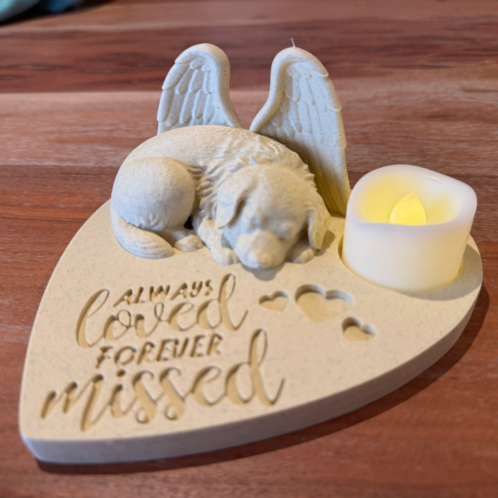 Memorial sculpture of a Great pyrenees with Angel wings wings on a heart-shaped base, featuring the text 'Always loved forever missed', with a small candle beside it on a wooden surface.
