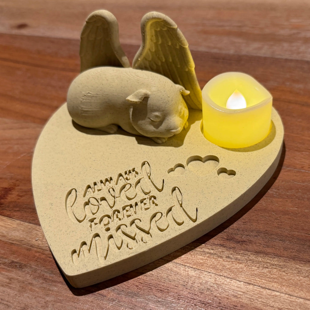 Heart-shaped plaque with a sleeping angel figurine of a Guinea pig and a lit candle on a wooden surface.
