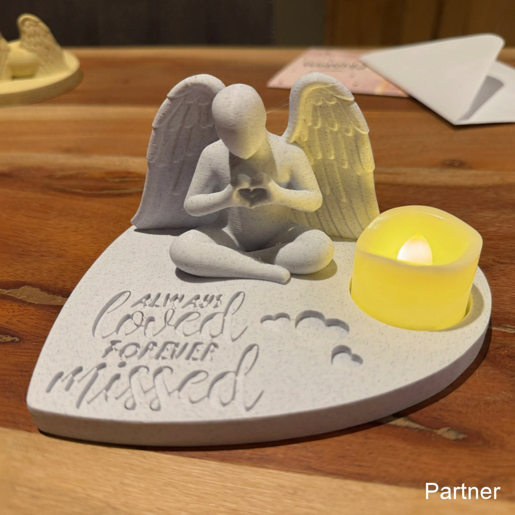 Memorial sculpture of an angel sitting on a heart-shaped base with a candle, on a wooden surface.