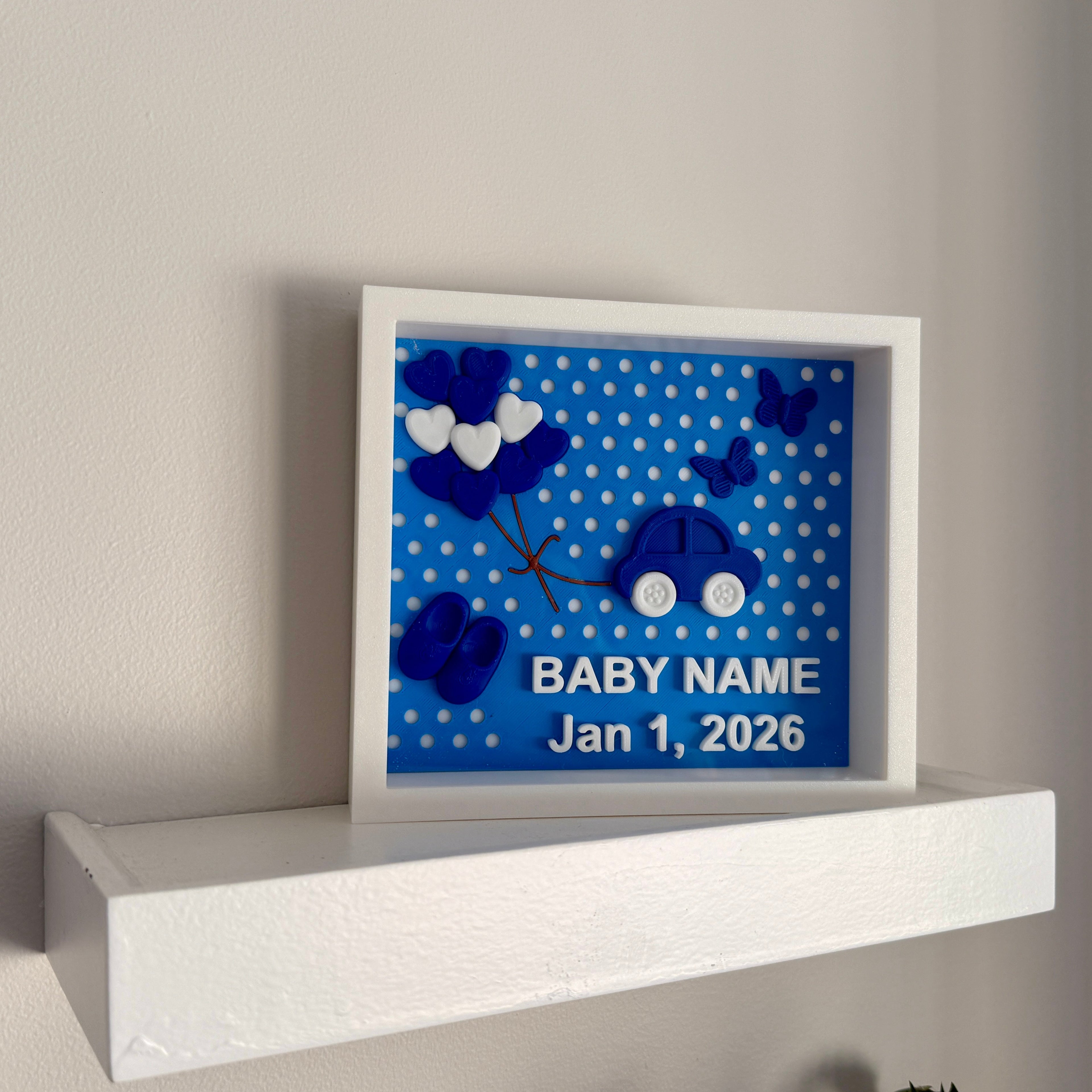 Framed baby milestone card with blue and white design on a white shelf against a beige wall.