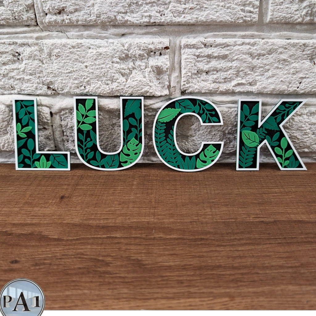 Decorative letters spelling 'LUCK' with leaf patterns against a stone wall and wooden surface.