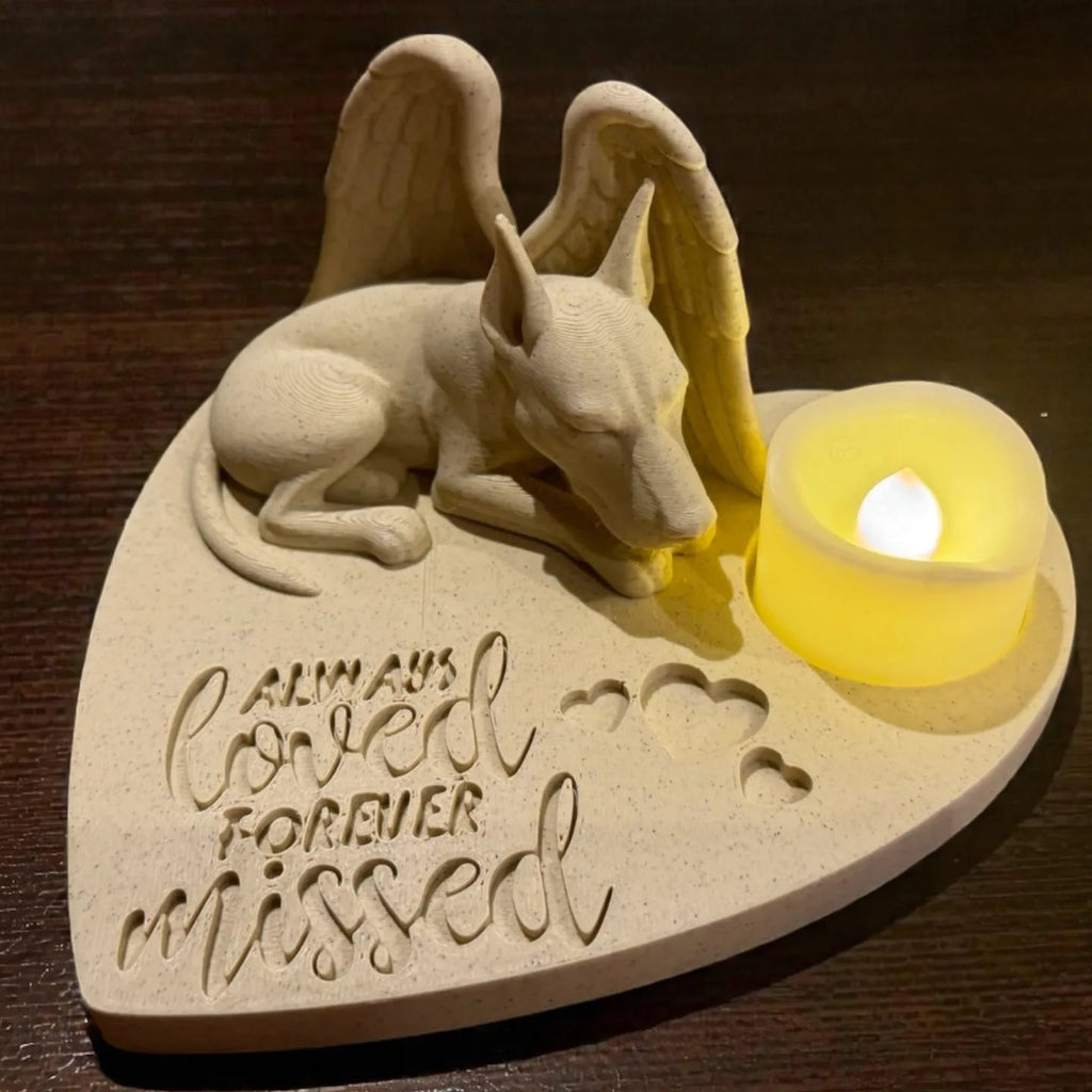 Memorial sculpture of a doberman with wings holding a candle, engraved with 'Always loved forever missed' on a wooden surface.
