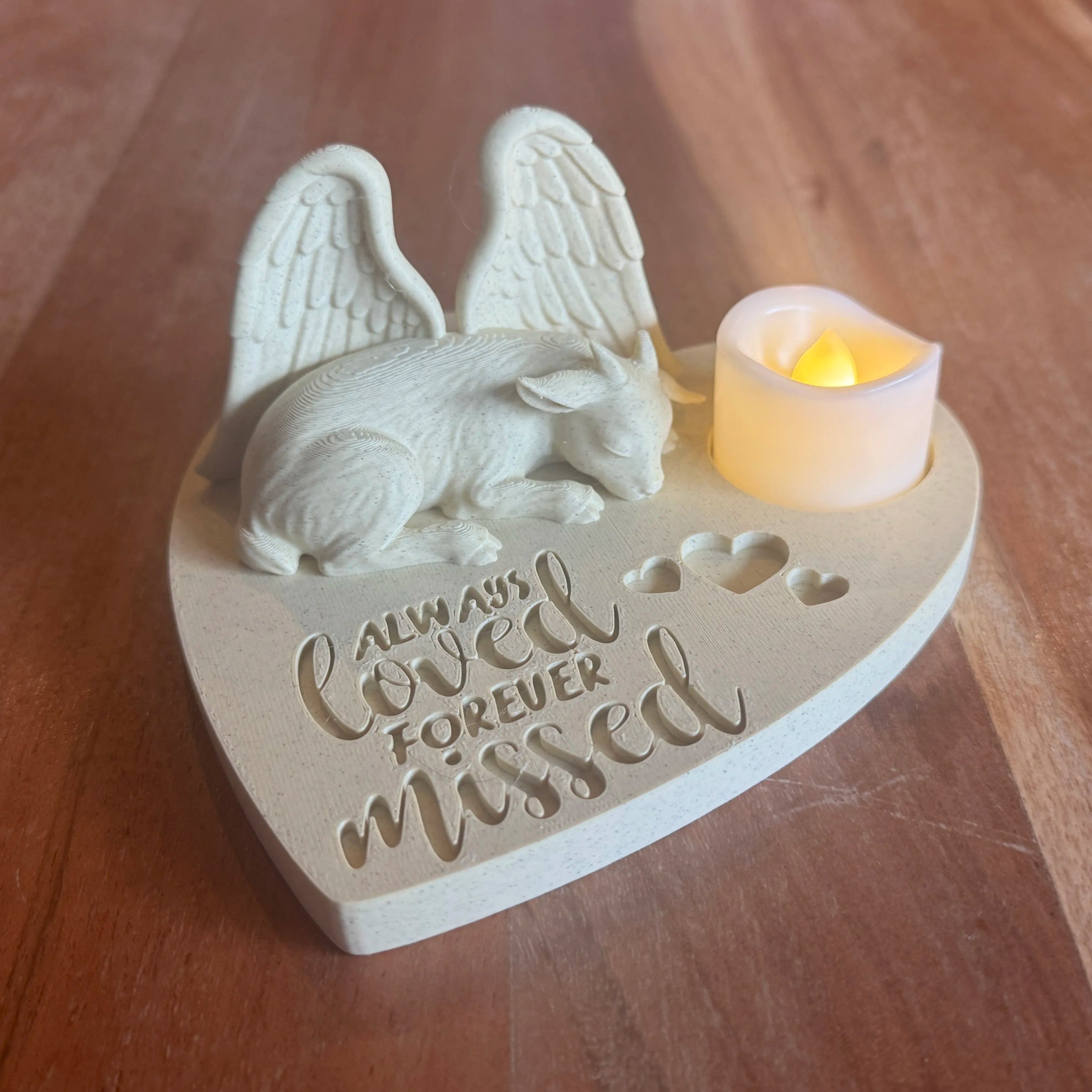 Heart-shaped memorial featuring a goat with angel wings and a lit candle on a wooden surface
