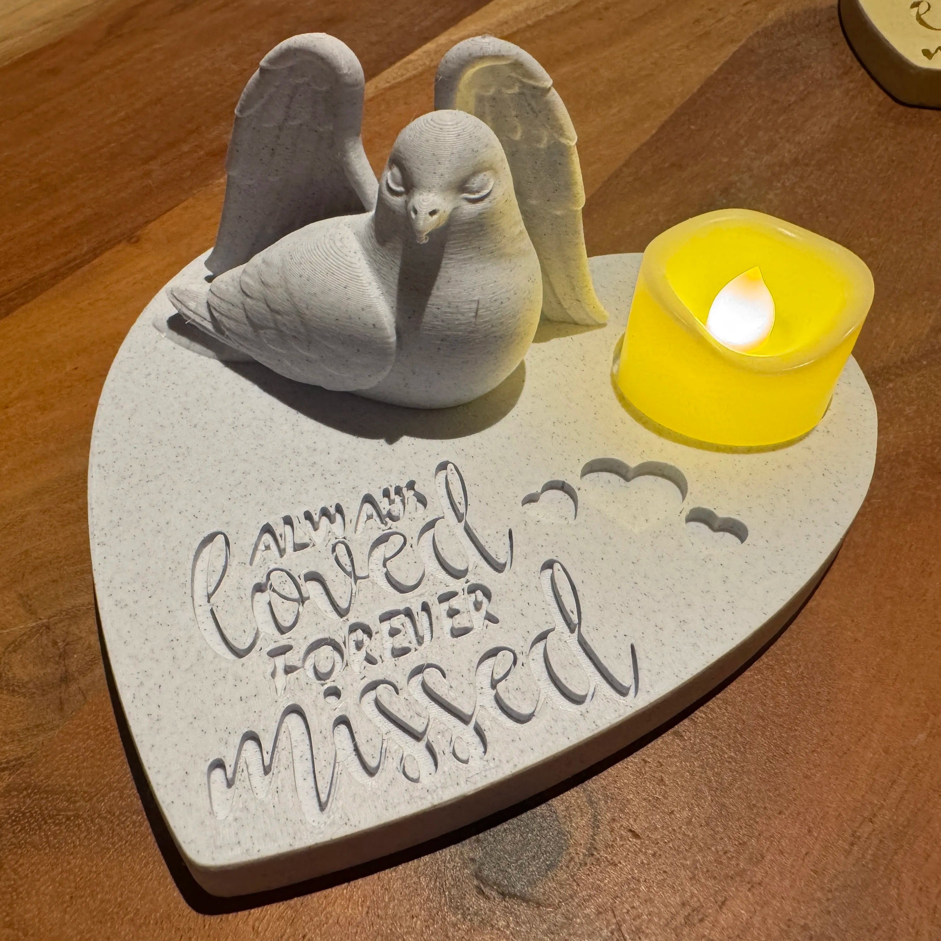 Memorial plaque with dove or pigeon wearing Angel wings and engraved text on a wooden surface with a small candle.