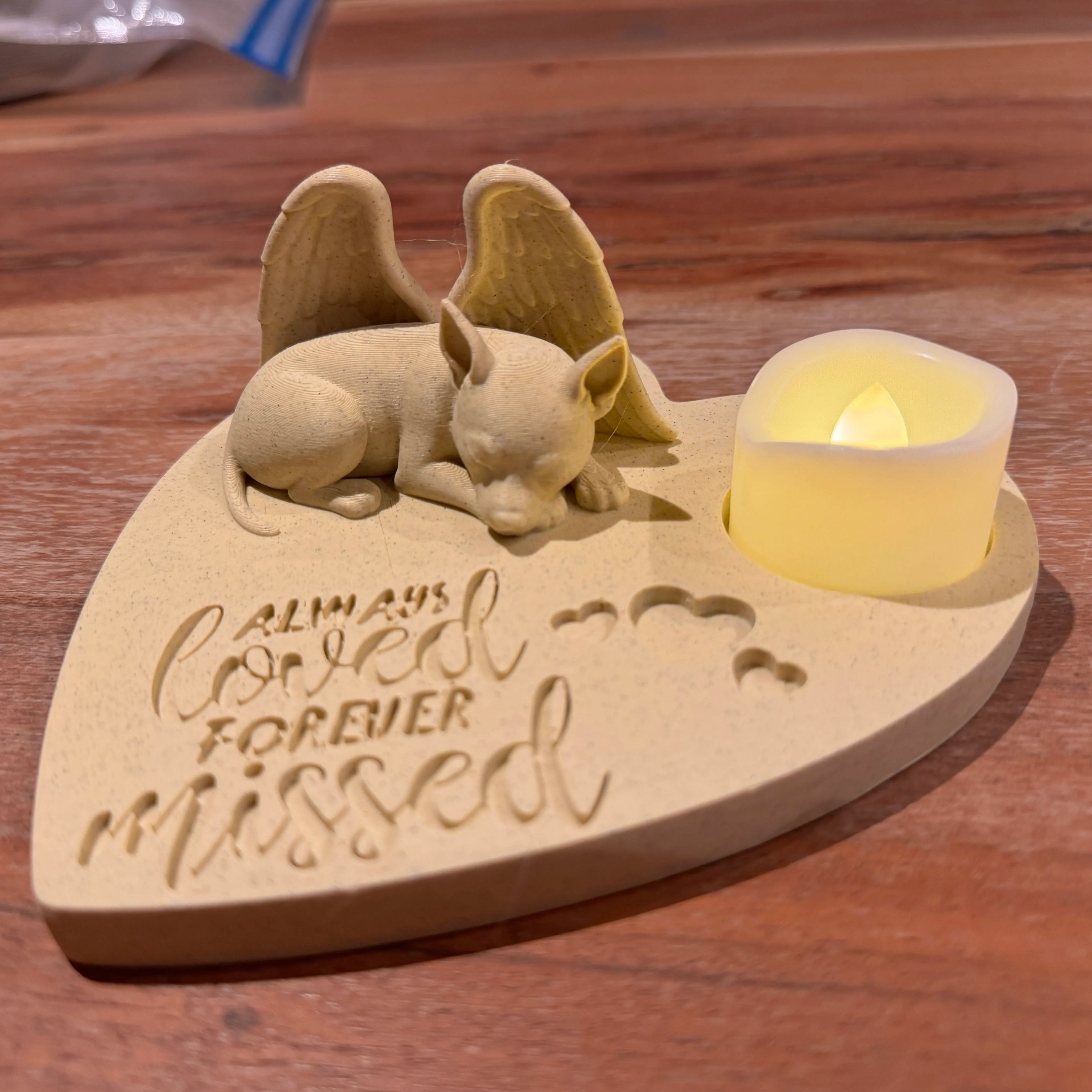 Heart-shaped wooden plaque with a pincer with wings figurine and a candle, engraved with 'Always loved forever missed', on a wooden surface.
