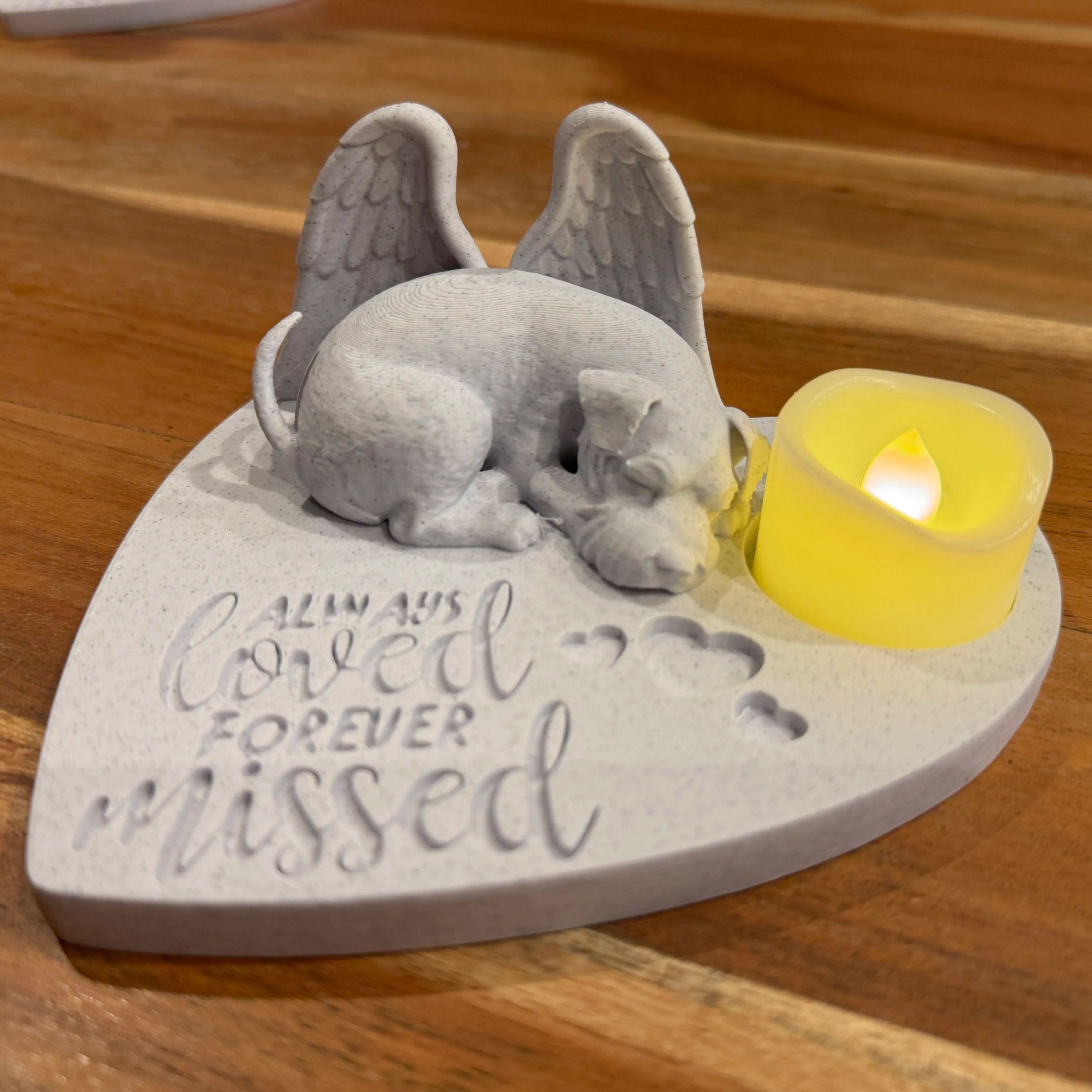 Memorial sculpture of a schnauzer with angel wings on a heart-shaped base with a candle, placed on a wooden surface.