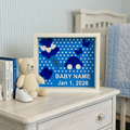 Framed baby milestone sign with blue polka dots and white text on a dresser with books and a teddy bear.
