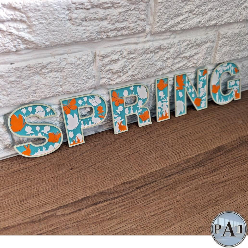 Decorative word 'SPRING' with floral patterns on a wooden surface against a stone wall.
