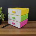 Stack of colorful drawers on a wooden surface with a plant and pens in the foreground.
