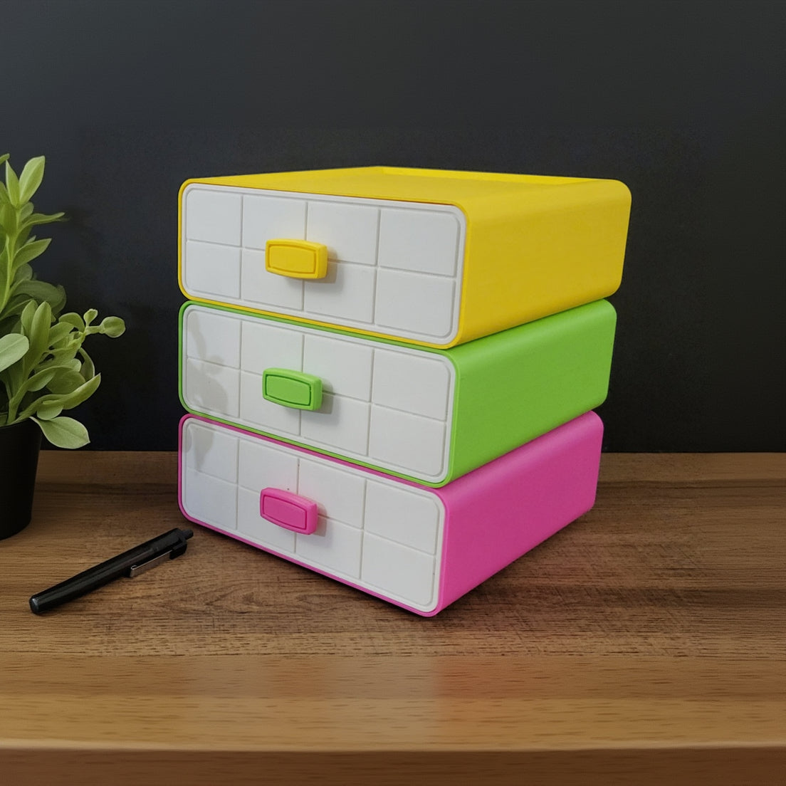 Stack of colorful drawers on a wooden surface with a plant and pens in the foreground.